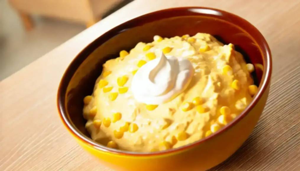 A bowl of creamy corn dip topped with a dollop of sour cream and sprinkled with sweet corn kernels on a wooden table.