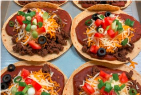 Homemade tortillas topped with seasoned beef, cheese, black olives, tomatoes, green onions, and refried beans on a baking sheet.