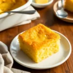 A golden slice of Easy Cornbread Casserole served on a white plate, with a buttery crust and moist texture, set on a rustic wooden table.