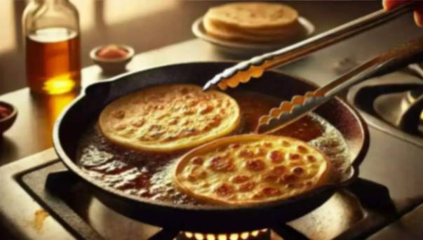 Two golden tortillas frying in oil in a skillet on a gas stovetop, with tongs flipping them and ingredients nearby.