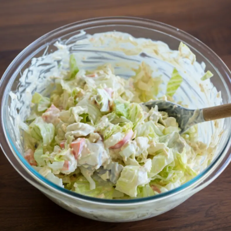 A bowl of fresh shredded lettuce, mayonnaise, diced red onions, chopped dill pickles, and banana peppers mixed in a clear glass bowl with a wooden spoon on a wooden surface.