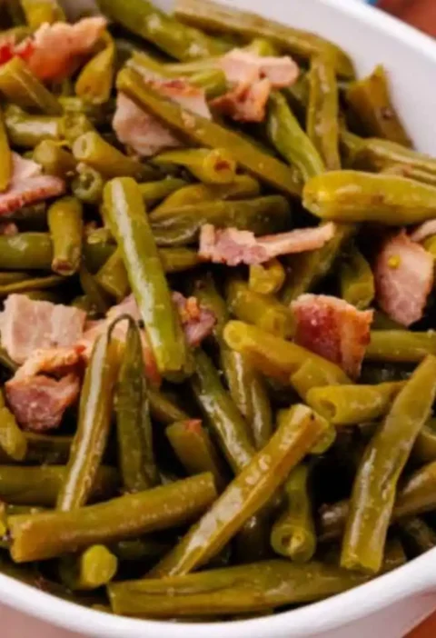 Cooked green beans mixed with tender bacon pieces served in a white bowl, showcasing a hearty and flavorful dish.