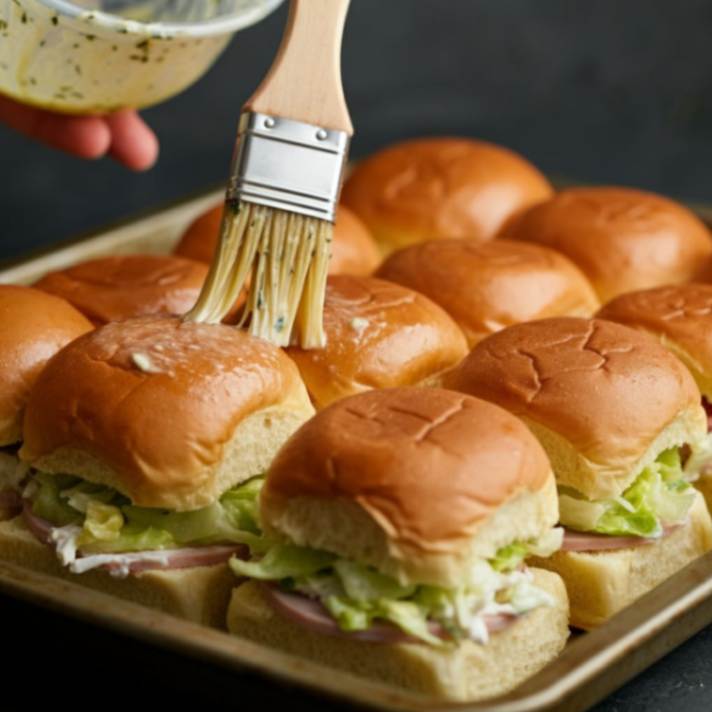 Brushing melted herb butter onto sliders with Deli meats and salad on soft golden buns placed on a baking tray.