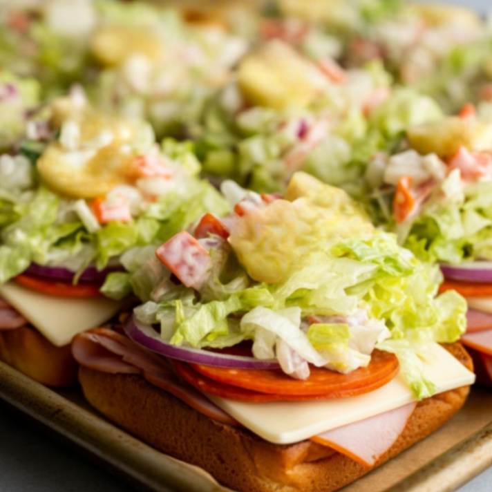 Open-faced sliders topped with Deli meats, cheese, lettuce salad, and dill pickles on a baking tray.