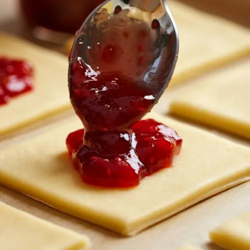 Spreading strawberry jam on dough squares with a spoon in the kitchen.