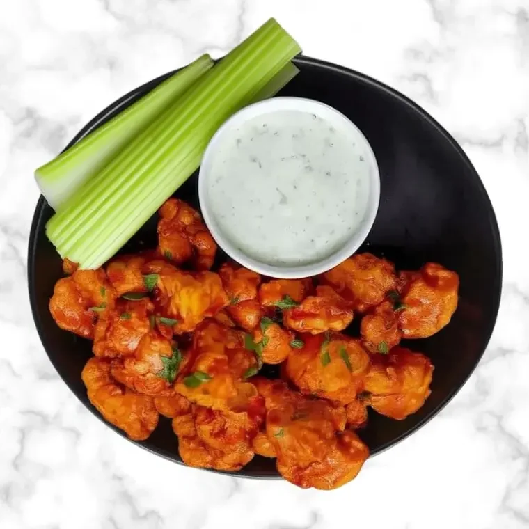 Buffalo cauliflower bites with ranch dip and celery sticks served in a black bowl on a marble background.