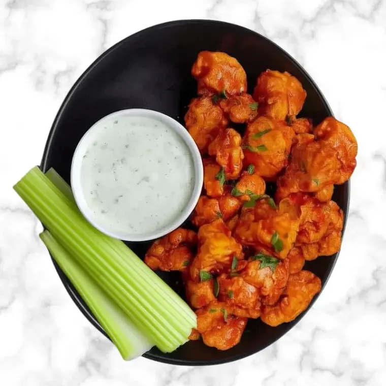 Buffalo cauliflower bites with ranch dip and celery sticks served in a black bowl on a marble background.
