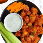 Air fryer buffalo cauliflower wings served with ranch dip, celery sticks, carrot sticks, and extra buffalo sauce in a black bowl.
