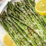 Air fryer asparagus spears generously topped with Parmesan cheese, chili flakes, and lemon slices served on a white ceramic plate.