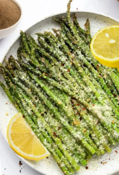 Air fryer asparagus spears generously topped with Parmesan cheese, chili flakes, and lemon slices served on a white ceramic plate.