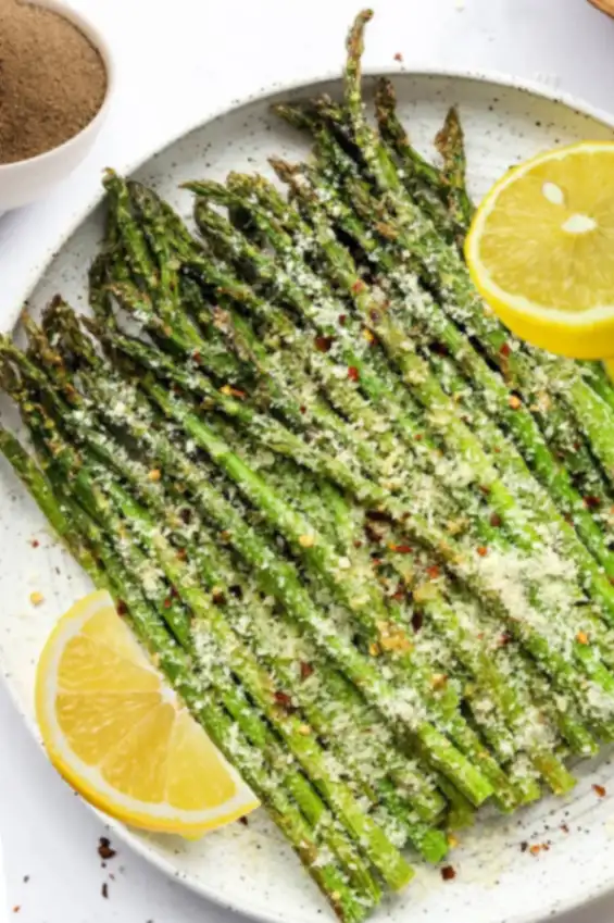 Air fryer asparagus spears generously topped with Parmesan cheese, chili flakes, and lemon slices served on a white ceramic plate.