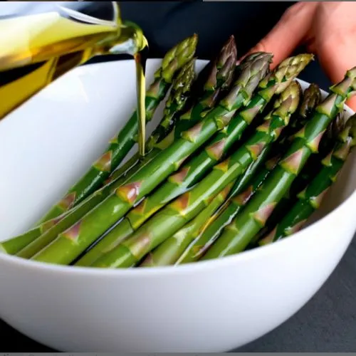 Fresh asparagus spears in a white bowl being drizzled with olive oil, prepared for air frying or cooking.
