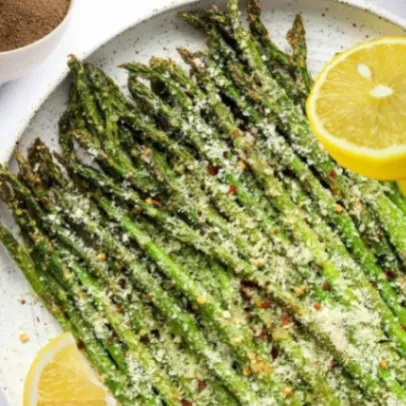 Air fryer asparagus spears topped with grated Parmesan cheese and red pepper flakes, served with lemon wedges on a plate.