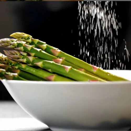 Fresh green asparagus in a white bowl being sprinkled with salt and black pepper, prepared for seasoning before cooking or air frying.