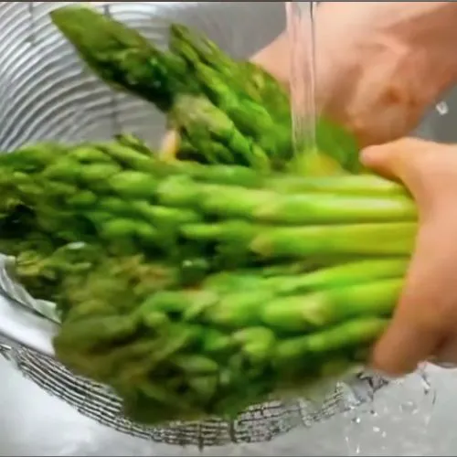 Person rinsing a bunch of fresh green asparagus under running water over a metal strainer in a kitchen sink.
