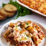 Plated cheesy rigatoni pasta with meat sauce, garnished with herbs and Parmesan, served with garlic bread and baked pasta dish.