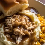 Mashed potatoes with shredded chicken and gravy, served with corn and a soft dinner roll on a white plate.