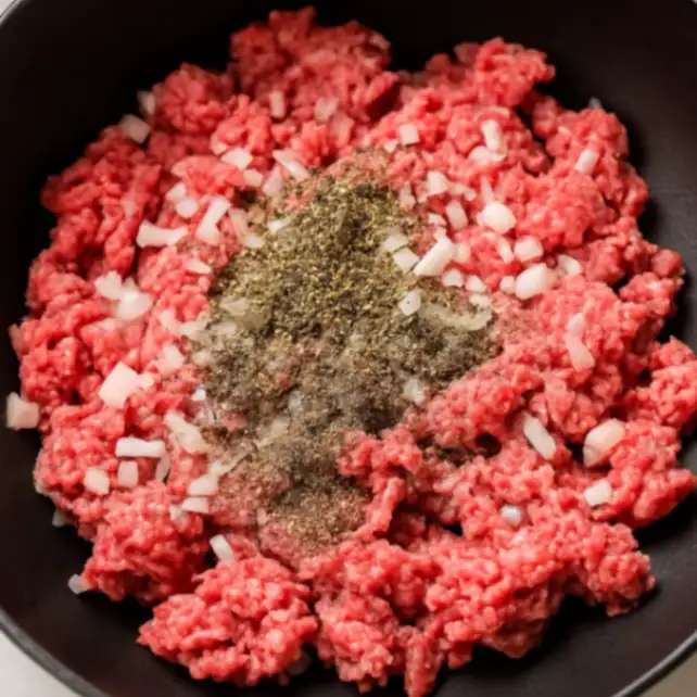 Ground beef in a skillet topped with diced onions and seasoning, ready to be cooked.
