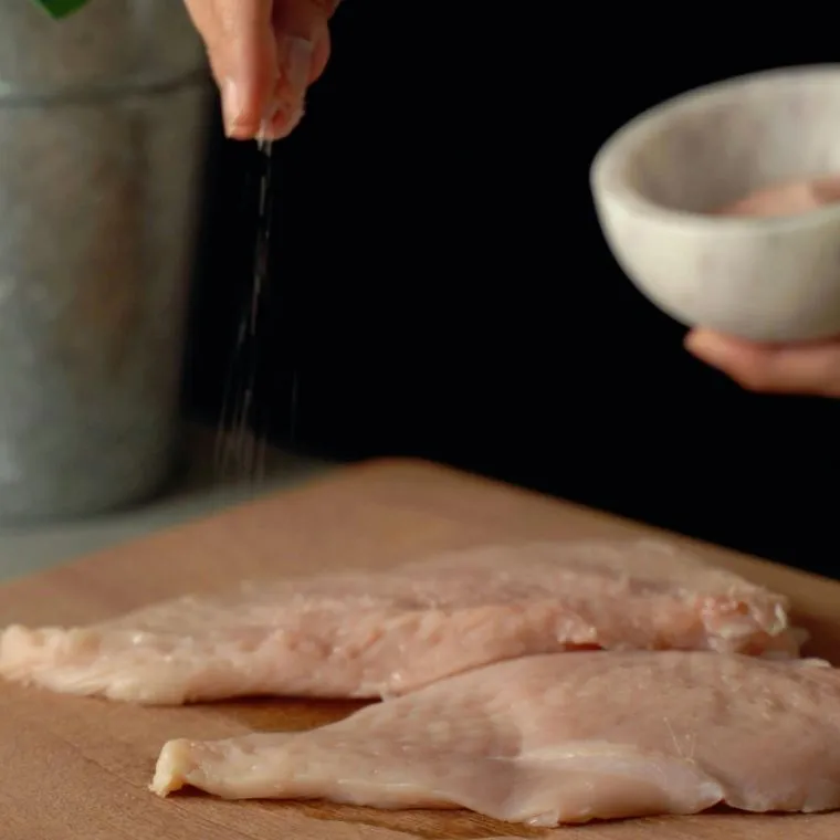 Hand seasoning raw chicken breasts with salt and pepper on a wooden cutting board, preparing them for cooking.